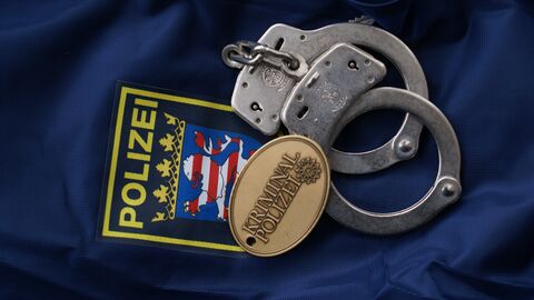 Auf einer blauen Decke liegen symbolisch Handfesseln und eine Kripomarke