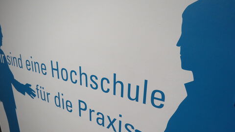 Eine Hochschule für die Praxis Blauer Schriftzug Wir sind eine Hochschule für die Praxis, rechts und links davon blaue Silhouetten von zwei Menschen