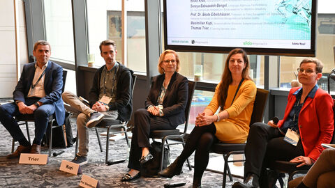 Teilnehmende Kongress 'Digitaler Staat 2024' sitzen in einer Diskussionsrunde, im Hintergrund ein eingeschalteter Monitor
