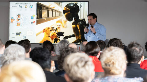  Herr Timo Paschke bei seinem Vortrag über Küstliche Intelligenz, im Vordergrund Publikum von hinten, im Hintergrund auf dem Beamer Hunde vor einem Roboter
