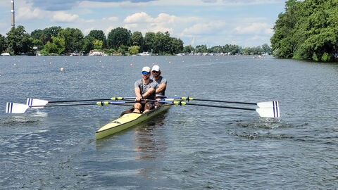 Das Bild zeigt einen See, im Vordergrund in Frontansicht Schäfer und Overath in ihrem Ruderboot