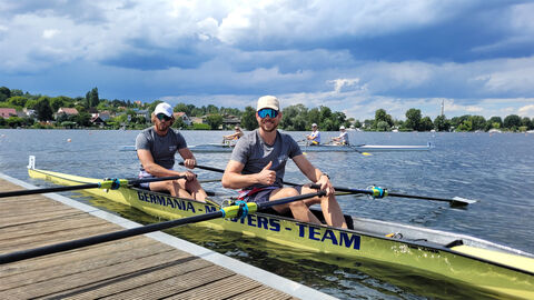 Schäfer und Overath sitzen in ihrem Boot an einer Anlegestelle und blicken in die Kamera, Overath sitzt vorne im Boot und reckt den Daumen