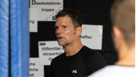 Auf dem Bild sieht man einen Trainer, der mit ernstem Ausdruck in Richtung einer Gruppe von Teilnehmern schaut. Die Gruppe hat an einem Blaulichtcamp teilgenommen, das im Kontext von Sicherheits- oder Rettungseinsätzen steht. Im Hintergrund sind verschiedene Hinweisschilder zu erkennen. Eines trägt die Aufschrift "Deeskalationstreppe", während andere Schilder mit Begriffen wie "Waffen" beschriftet sind. Die Szenerie wirkt sachlich und konzentriert, als ob eine wichtige Lektion in Konfliktbewältigung oder Gewaltprävention vermittelt wird. Auf dem Bild sieht man einen Trainer, der mit ernstem Ausdruck in Richtung einer Gruppe von Teilnehmern schaut. Die Gruppe hat an einem Blaulichtcamp teilgenommen, das im Kontext von Sicherheits- oder Rettungseinsätzen steht. Im Hintergrund sind verschiedene Hinweisschilder zu erkennen. Eines trägt die Aufschrift "Deeskalationstreppe", während andere Schilder mit Begriffen wie "Waffen" beschriftet sind. Die Szenerie wirkt sachlich und konzentriert, als ob eine wichtige Lektion in Konfliktbewältigung