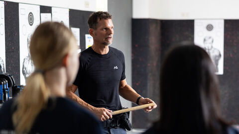 Auf dem Bild ist ein Einsatztrainer zu sehen, der konzentriert zu einer Gruppe spricht. In seiner Hand hält er einen langen Holzstab, der für das Training verwendet wird. Im Vordergrund stehen mehrere weibliche Teilnehmerinnen, die aufmerksam zuhören. Sie tragen T-Shirts mit der Aufschrift "Blaulichtcamp". Die Szene wirkt wie eine praktische Übung, bei der der Trainer den Teilnehmerinnen den Umgang mit Ausrüstung oder Techniken für den Einsatz vermittelt. Auf dem Bild ist ein Einsatztrainer zu sehen, der konzentriert zu einer Gruppe spricht. In seiner Hand hält er einen langen Holzstab, der für das Training verwendet wird. Im Vordergrund stehen mehrere weibliche Teilnehmerinnen, die aufmerksam zuhören. Sie tragen T-Shirts mit der Aufschrift "Blaulichtcamp". Die Szene wirkt wie eine praktische Übung, bei der der Trainer den Teilnehmerinnen den Umgang mit Ausrüstung oder Techniken für den Einsatz vermittelt.