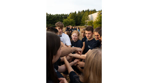 Das Bild zeigt die gleiche Gruppe von Teilnehmern des Blaulichtcamps, dieses Mal aus einer anderen Perspektive. Die Gruppe steht weiterhin auf dem Sportplatz und spielt ein kommunikatives Gruppenspiel. Durch den veränderten Blickwinkel ist die Anordnung der Teilnehmer etwas anders zu sehen, doch die spielerische und gemeinschaftliche Atmosphäre bleibt bestehen. Man erkennt, dass das Spiel darauf abzielt, die Interaktion und das Teamgefühl zu fördern. Das Bild zeigt die gleiche Gruppe von Teilnehmern des Blaulichtcamps, dieses Mal aus einer anderen Perspektive. Die Gruppe steht weiterhin auf dem Sportplatz und spielt ein kommunikatives Gruppenspiel. Durch den veränderten Blickwinkel ist die Anordnung der Teilnehmer etwas anders zu sehen, doch die spielerische und gemeinschaftliche Atmosphäre bleibt bestehen. Man erkennt, dass das Spiel darauf abzielt, die Interaktion und das Teamgefühl zu fördern.