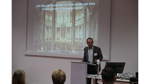 Reiner Beck Vorsitzender Zentrum Demokratieforschung Hessen steht vor einem Rednerpult, Im Hintergrund eine Präsentation mit dem Titel 'Der Deutsche Weg zur Demokratie war alles andere als eine Erfolgsgeschichte