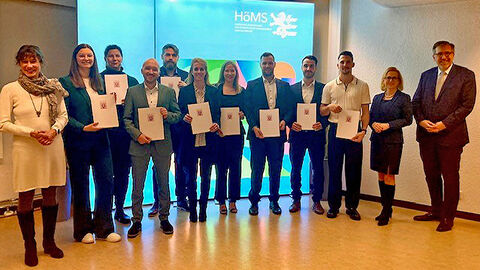 Absolventinnen und Absolventen stehen im Halbkreis zusammen, blicken in die Kamera und halten hr Abschlusszeugnis in der Hand