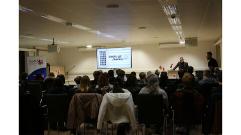 Das Bild zeigt einen Saal mit Blick auf die Dozierenden. Im Vordergrund sind Köpfe der zuhörenden zu erkennen. Auf der Leinwand an der Wand ist eine Powerpoint-Folie mit dem Schriftzug "Hanau ist überall" zu erkennen.