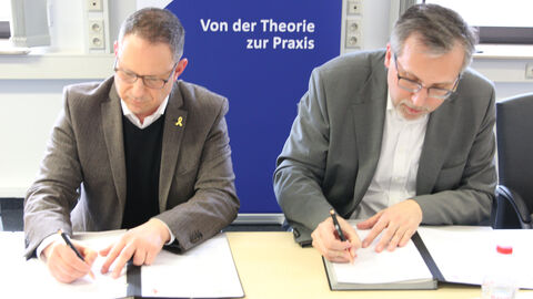 Steve Landau und Dr. Walter Seubert sitzen an einem Tisch und unterzeichnen jeweils die Kooperationsvereinbarung