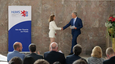 Innenstaatssekretär Martin Rößler reicht vor Publikum einer jungen Frau die Hand und hält ein Blatt in der Hand.