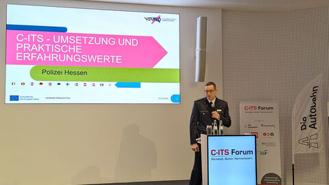 Das Foto zeigt Ulf Mielinger in Uniform an einem Pult mit der Aufschrift C-ITS Forum. Er hält ein Mikrofon in der Hand und trägt offensichtlich vor. Im Hintergrund ist eine große Leinwand zu erkennen. Dort ist eine Präsentation gezeigt, die im oberen Bildrand das Logo des Projektes VERKKO trägt. In der Mitte der Präsentation ist die Aufschrift C-ITS Umsetzung und praktische Erfahrungswerte zu lesen. Darunter steht Polizei Hessen. Ebenfalls darunter befinden sich 12 Länderflaggen in kreisrunden Darstellung. 